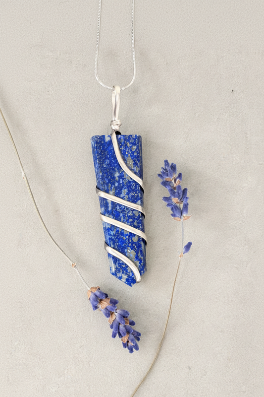 Lapis lazuli wire wrapped pendant showing deep cobalt blue with golden pyrite flecks on stainless steel snake chain, styled with dried lavender